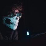 a person with a skull mask reviews code that’s reflected in their glasses.
