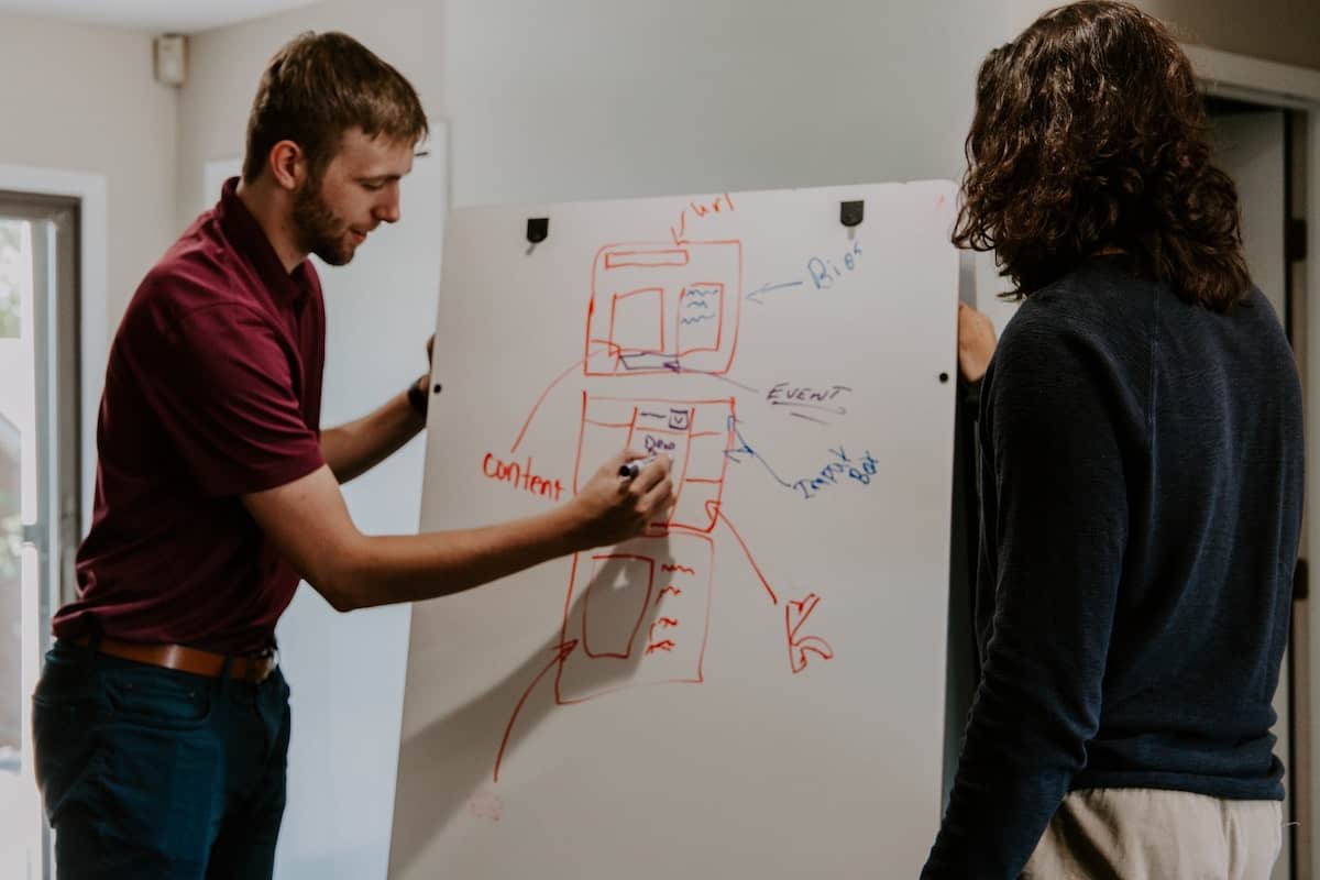 A man draws a diagram of a data structure on a dry erase board as his colleague looks on.