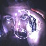 A bearded man in a dark room holds up a bundle of bright string lights to his face.