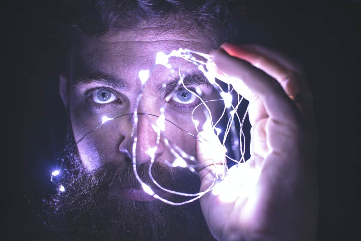 A bearded man in a dark room holds up a bundle of bright string lights to his face.