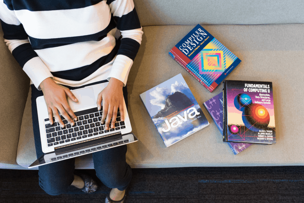 A woman with a laptop sits beside several books on web design, computer science, and Java programming women in stem