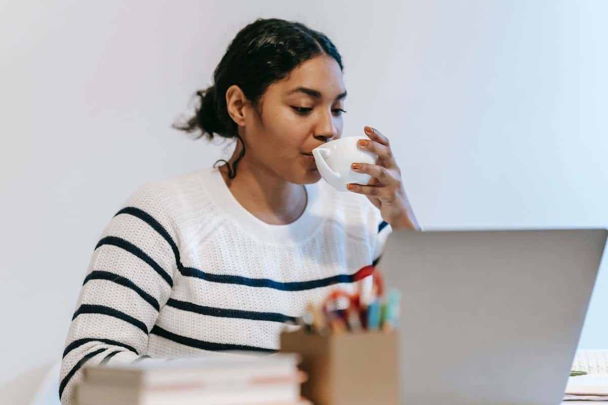 : a young woman working on a laptop while drinking out of a white cup coding bootcamp cost