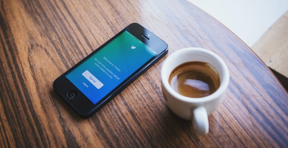 A mobile phone flashing the Twitter app and a cup of coffee sit side-by-side on a table. Tech Jobs at Twitter: Requirements and Job Description