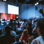 Photo of an audience of attendees at an event looking at two big white screens